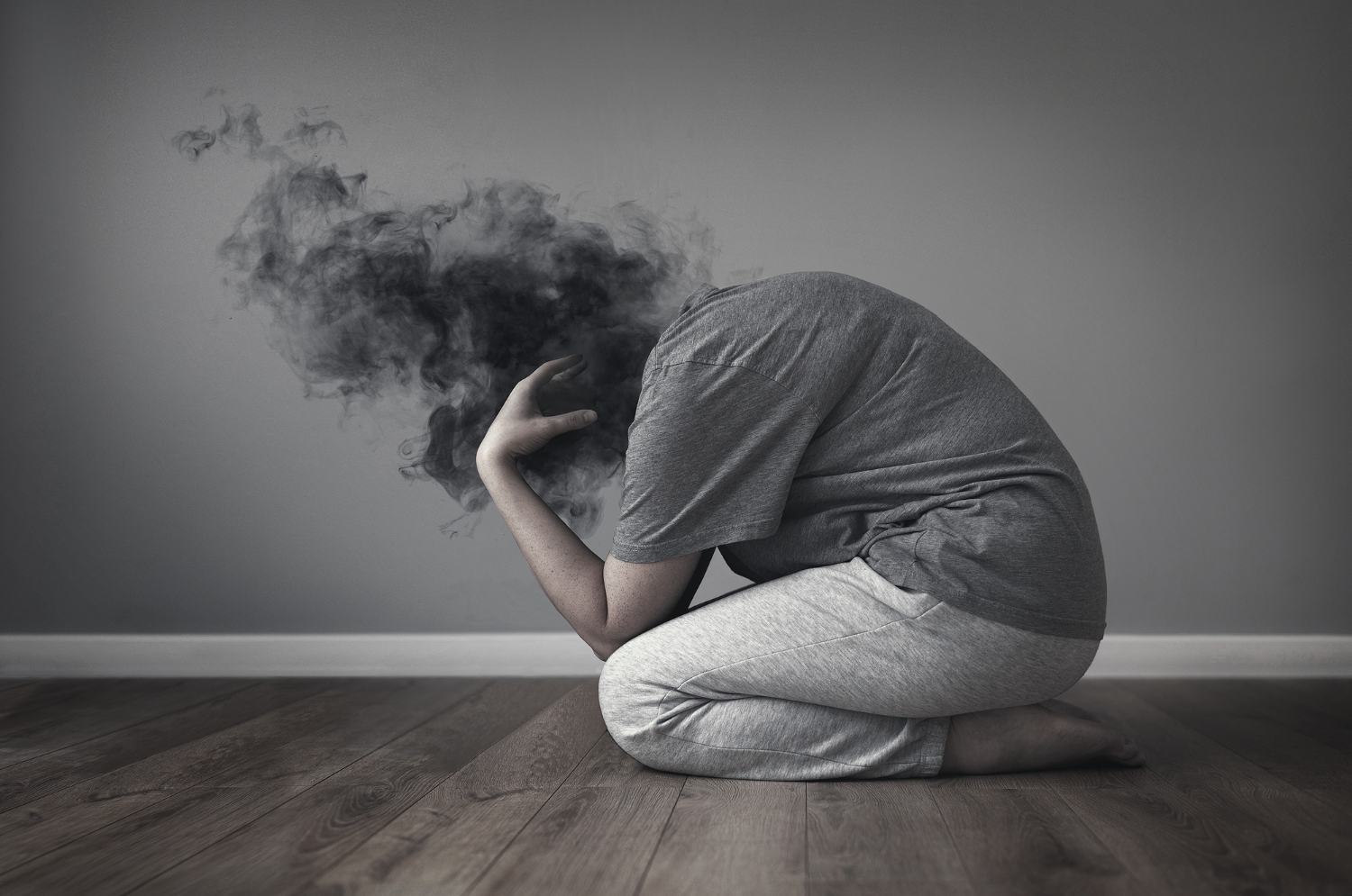 Photographie en noir et blanc - Une femme vêtue d'un survêtement et d'un tee-shirt est agenouillée sur un parquet seule - sa tête n'est pas visible, une fumée noire se trouve à sa place - Dépression et perte de soi